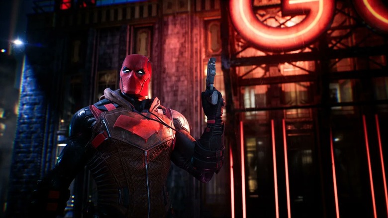 A screenshot from Gotham Knights, showing Red Hood standing in front of a building which is lit up red. A pistol is in his left hand.