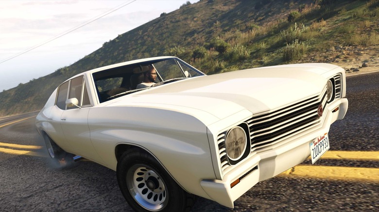 A screenshot from GTA Online showing a character driving a white muscle car down a road.